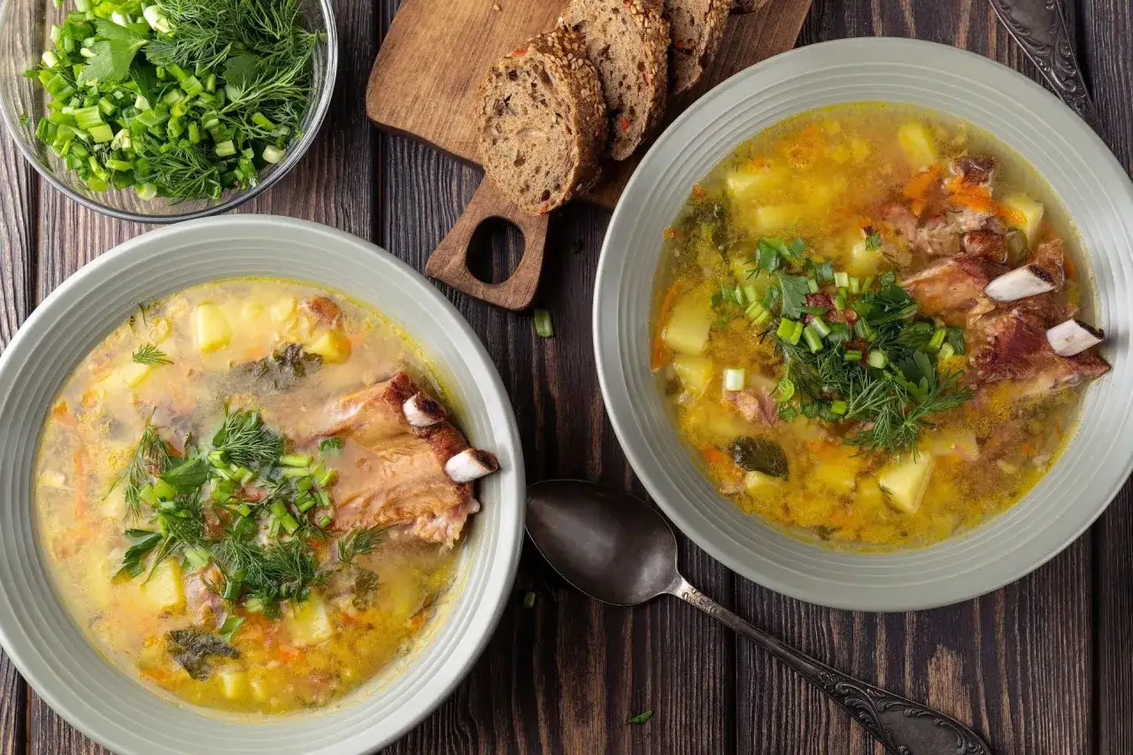 Dwie miski zupy grochowej z mięsem, ziemniakami i ziołami, obok chleb i miska z posiekaną zieleniną.