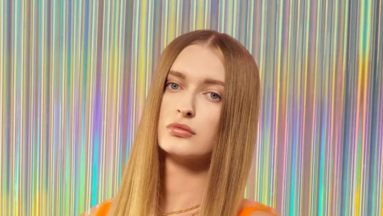 Sophie, top model o długich, blond włosach, pozuje na tle błyszczącej, tęczowej kurtyny.