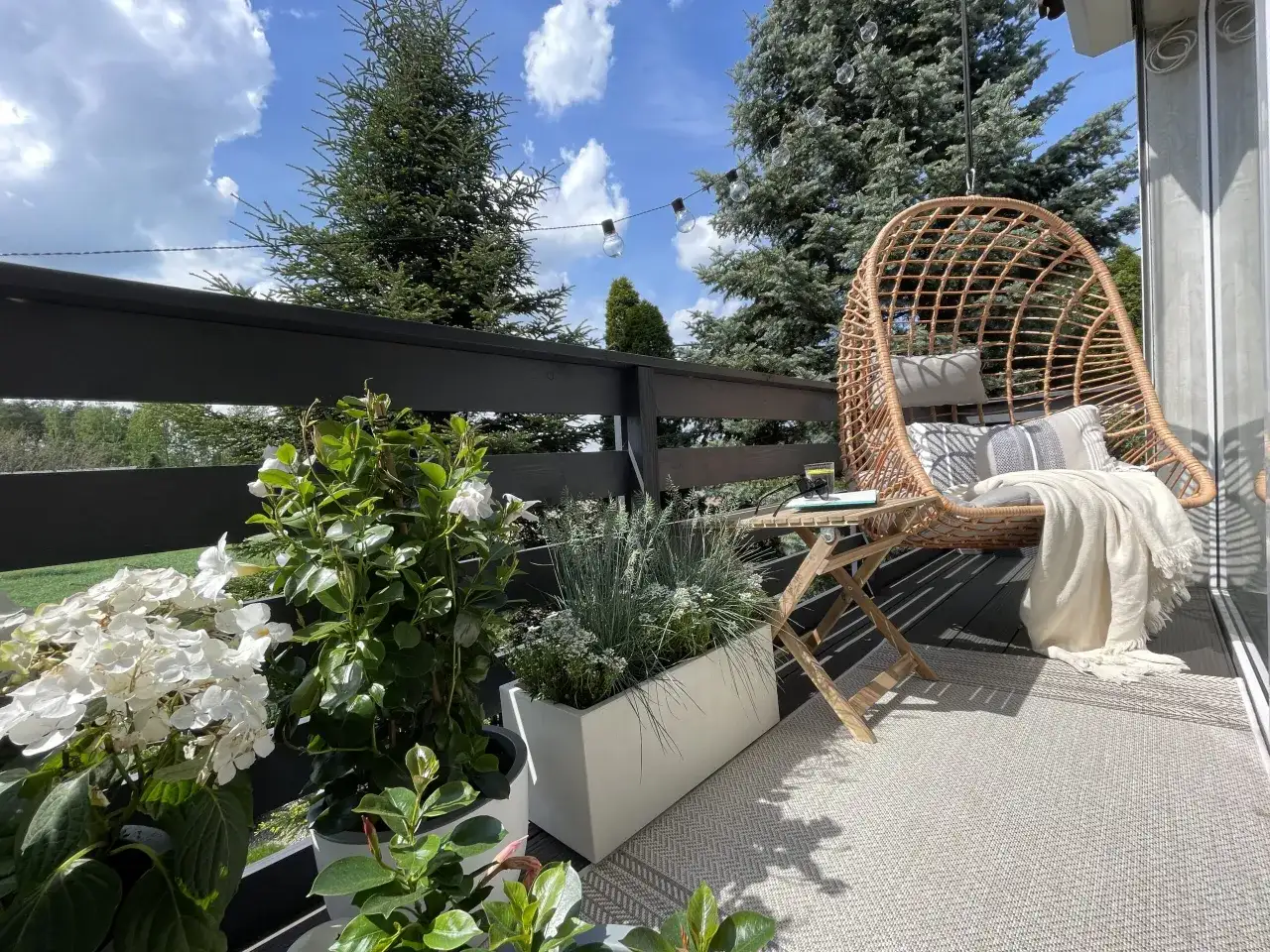 Przytulny mini balkon w bloku z fotelem wiszącym, stolikiem i kwitnącymi roślinami. Idealne miejsce na relaks.