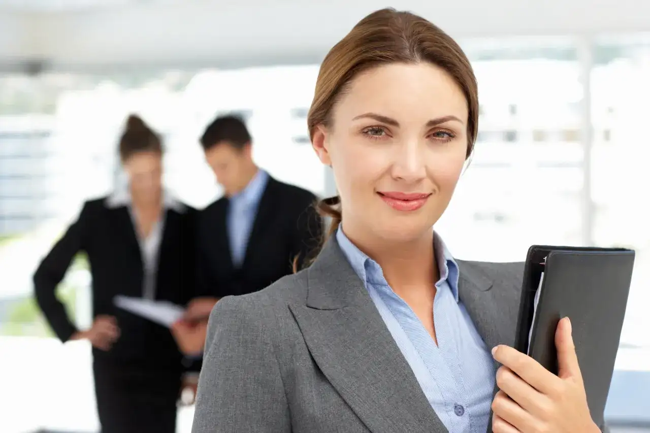 Uśmiechnięta bizneswoman z teczką, w tle współpracownicy omawiający dokumenty. Widać, że panuje między nimi umowa dorozumiana.