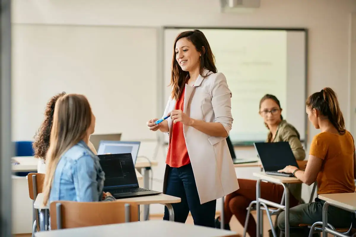 Nauczycielka wyjaśnia studentom, jak zostać nauczycielem po innych studiach. Uczniowie pracują na laptopach w sali lekcyjnej.