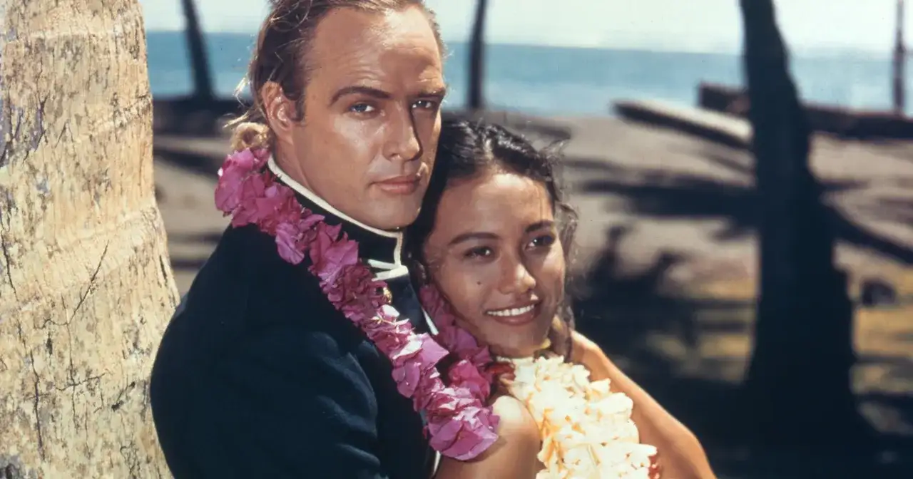 Marlon Brando z kobietą w kwiatowych girlandach na tle palmy i oceanu.