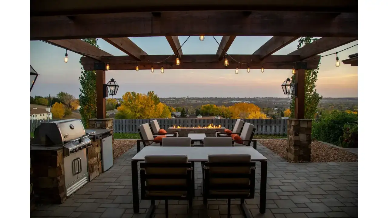 Oświetlenie tarasu inspiracje: przytulny kącik z grillem, stołem i paleniskiem pod drewnianą pergolą, z widokiem na jesienny krajobraz.