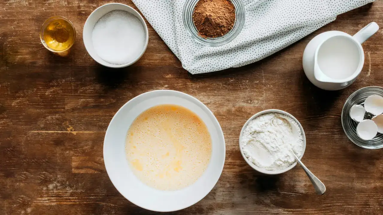 Składniki do pieczenia na drewnianym blacie: jajka, cukier, mąka, mleko, kakao, miód.