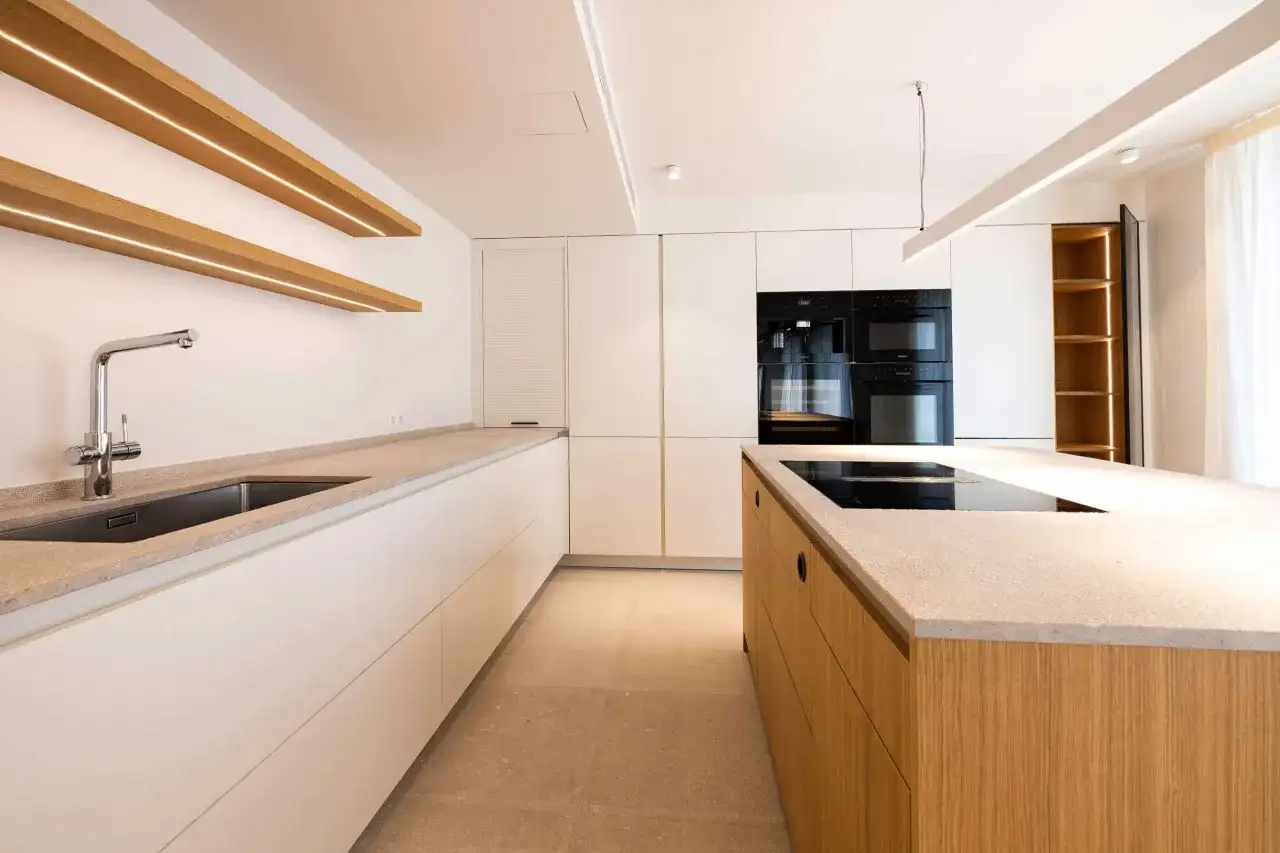 Nowoczesna kuchnia z wyspą i wbudowanymi urządzeniami. Ciepłe, drewniane półki i oświetlenie cokołu w kuchni tworzą przytulną atmosferę.