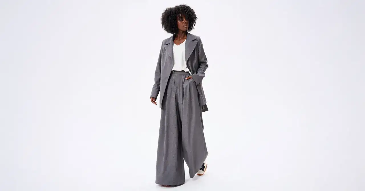 Kobieta w szarym garniturze z szerokimi spodniami i marynarką, stojąca na białym tle.