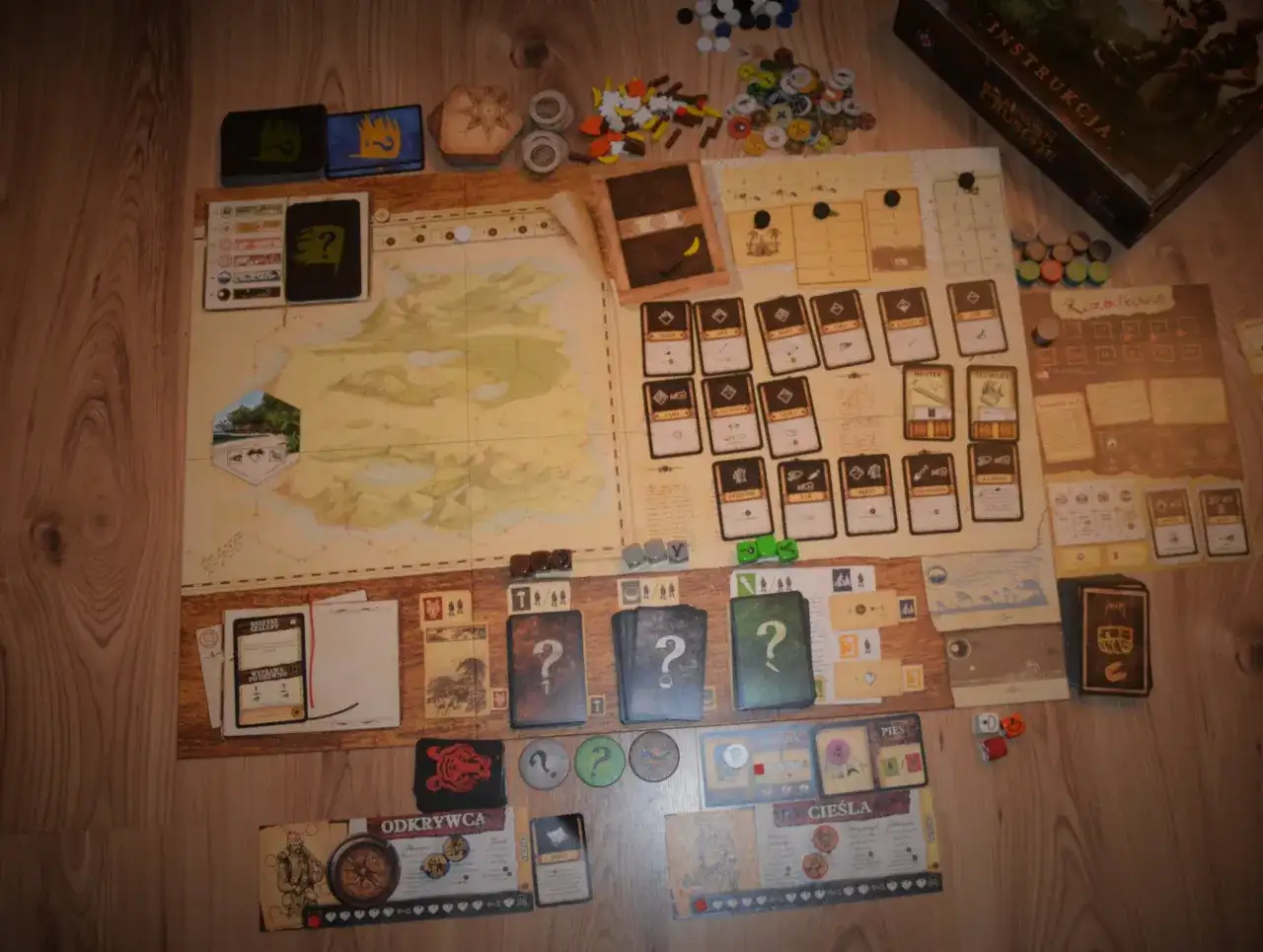 Plansza gry planszowej "Robinson Crusoe" z kartami, żetonami i instrukcją. Recenzja książki Robinson Crusoe w tle.