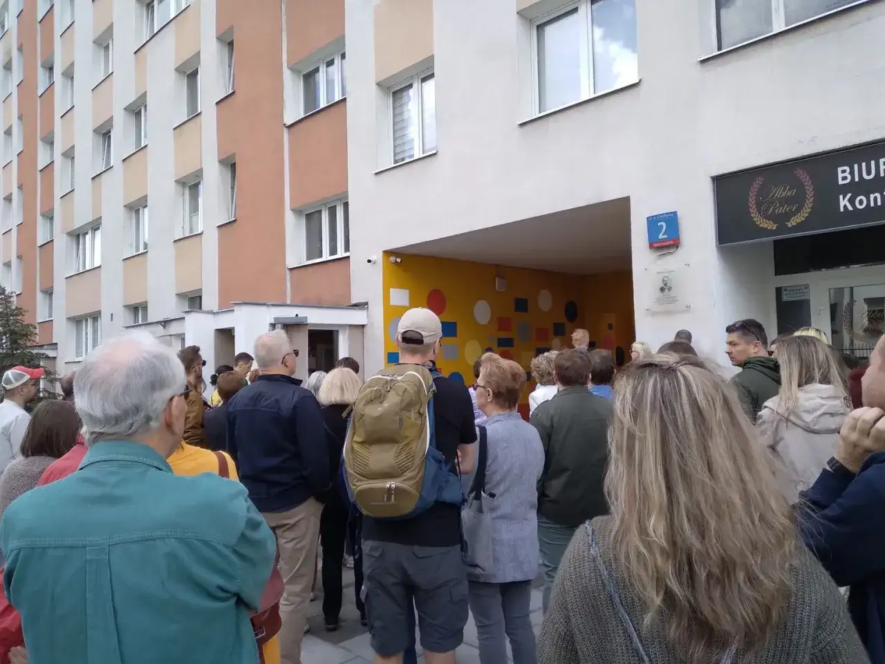 Grupa ludzi stoi przed blokiem mieszkalnym z kolorową ścianą i numerem 2.