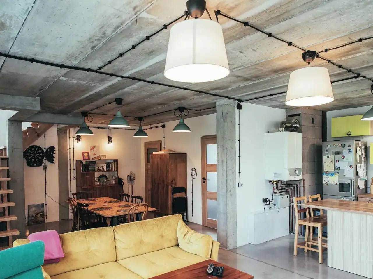 Nowoczesne wnętrze z betonowym sufitem i widoczną instalacją natynkową czarną. Wygodna, żółta kanapa i jadalnia tworzą przytulną przestrzeń.