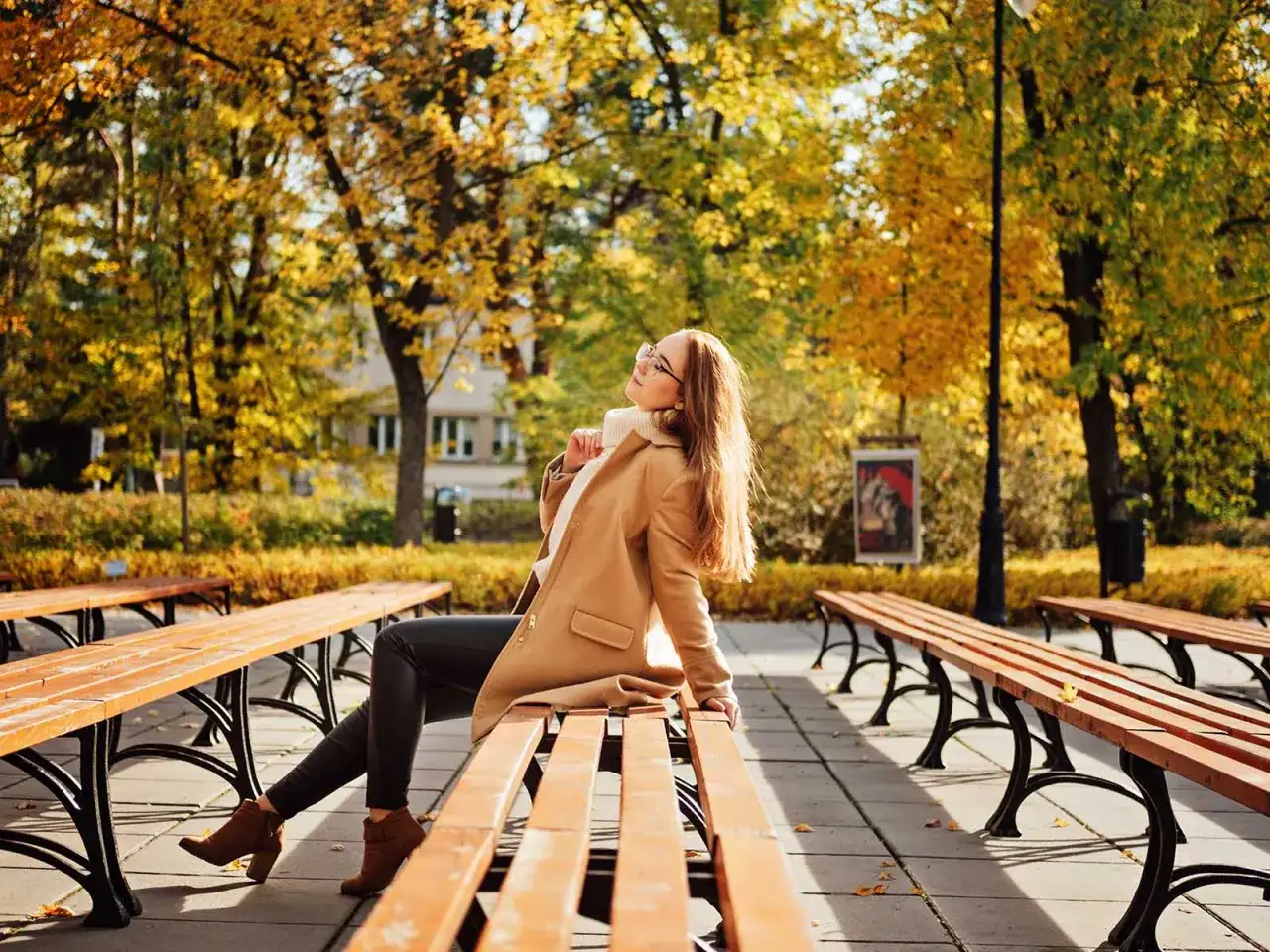 Kobieta siedzi na ławce w parku, otoczona jesiennymi barwami. Jej twarz skierowana ku słońcu, emanuje spokojem i pozytywnymi emocjami.