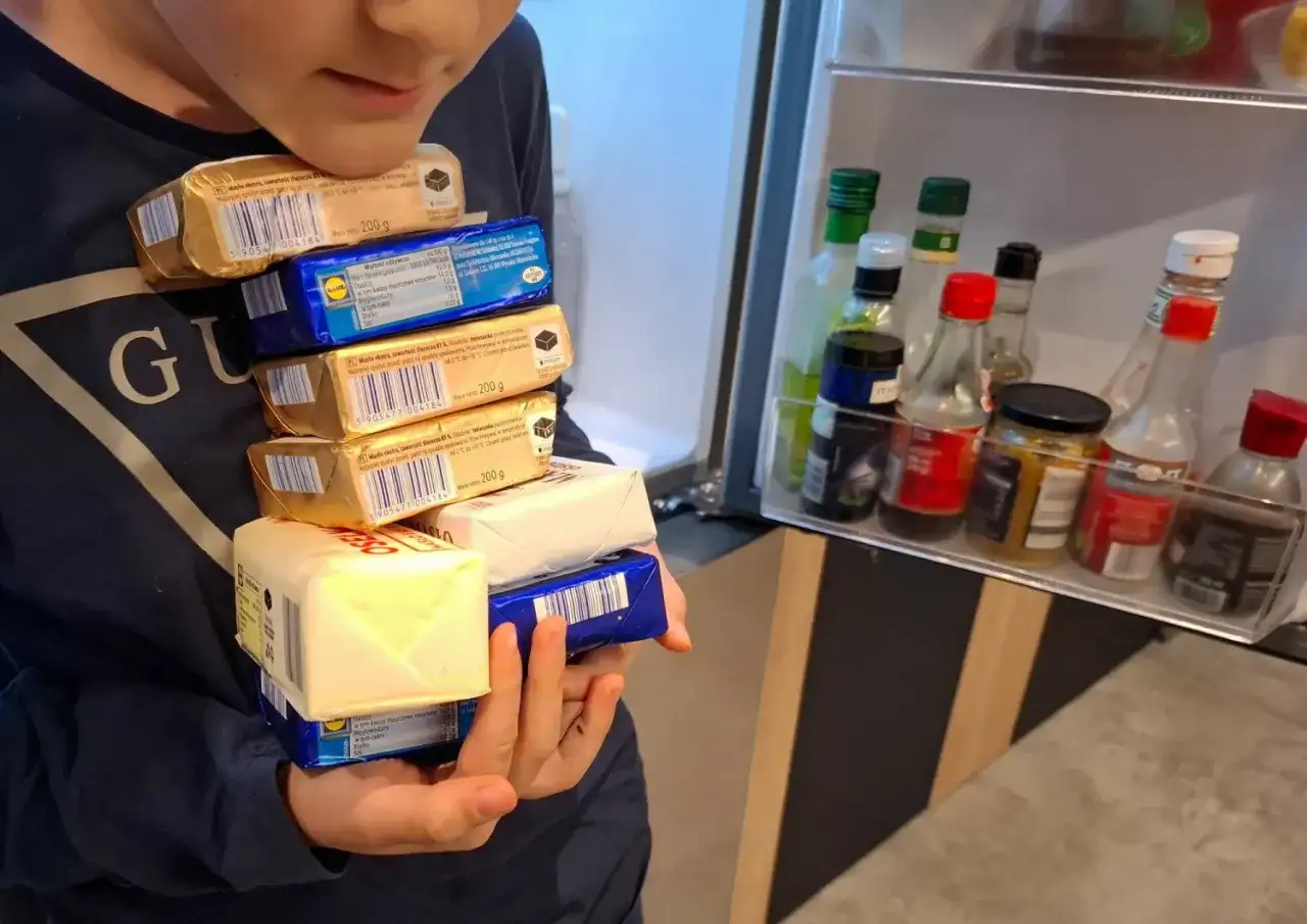 Dziecko trzyma stos opakowań masła przed otwartą lodówką z butelkami sosów.
