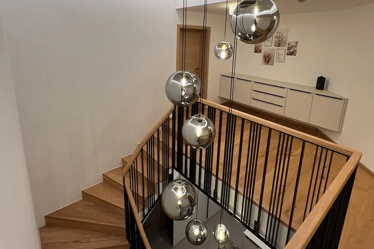 Nowoczesna klatka schodowa z drewnianymi stopniami i czarną balustradą, ozdobiona wiszącymi kulistymi lampami.