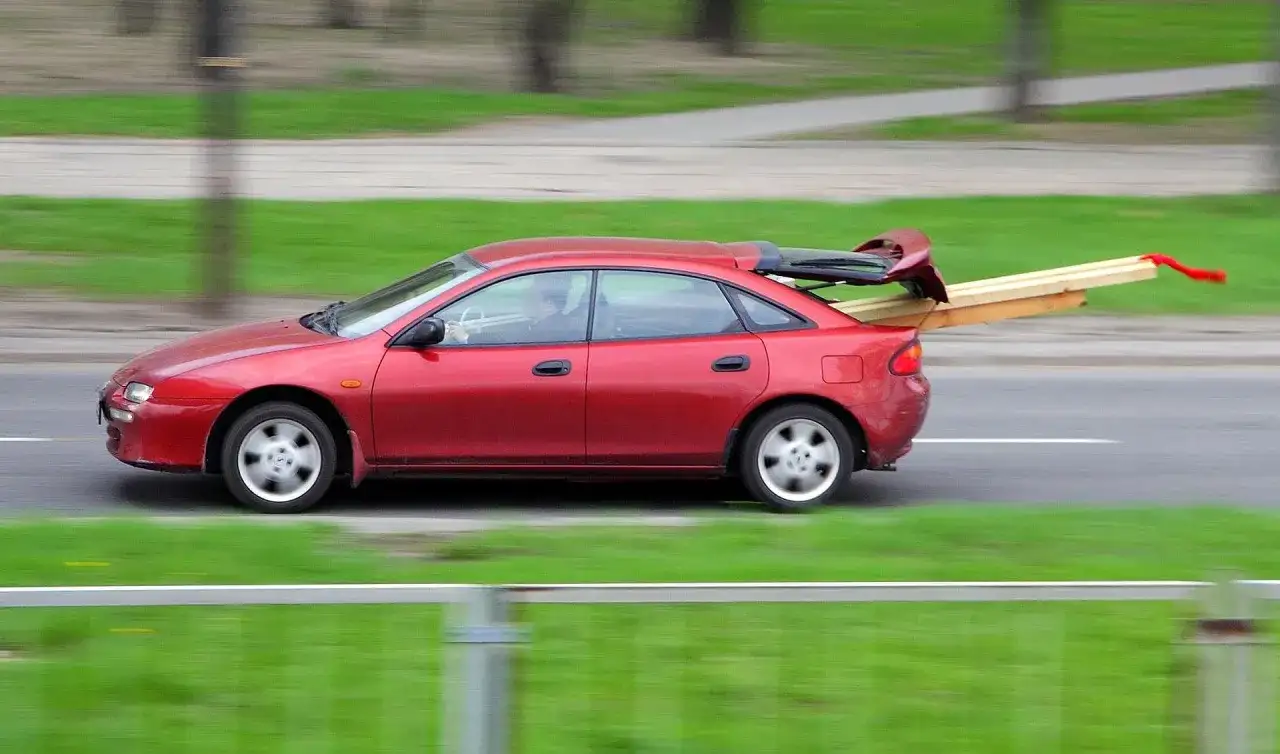 Czerwony sedan jedzie z otwartym bagażnikiem, w którym przewozi deski.