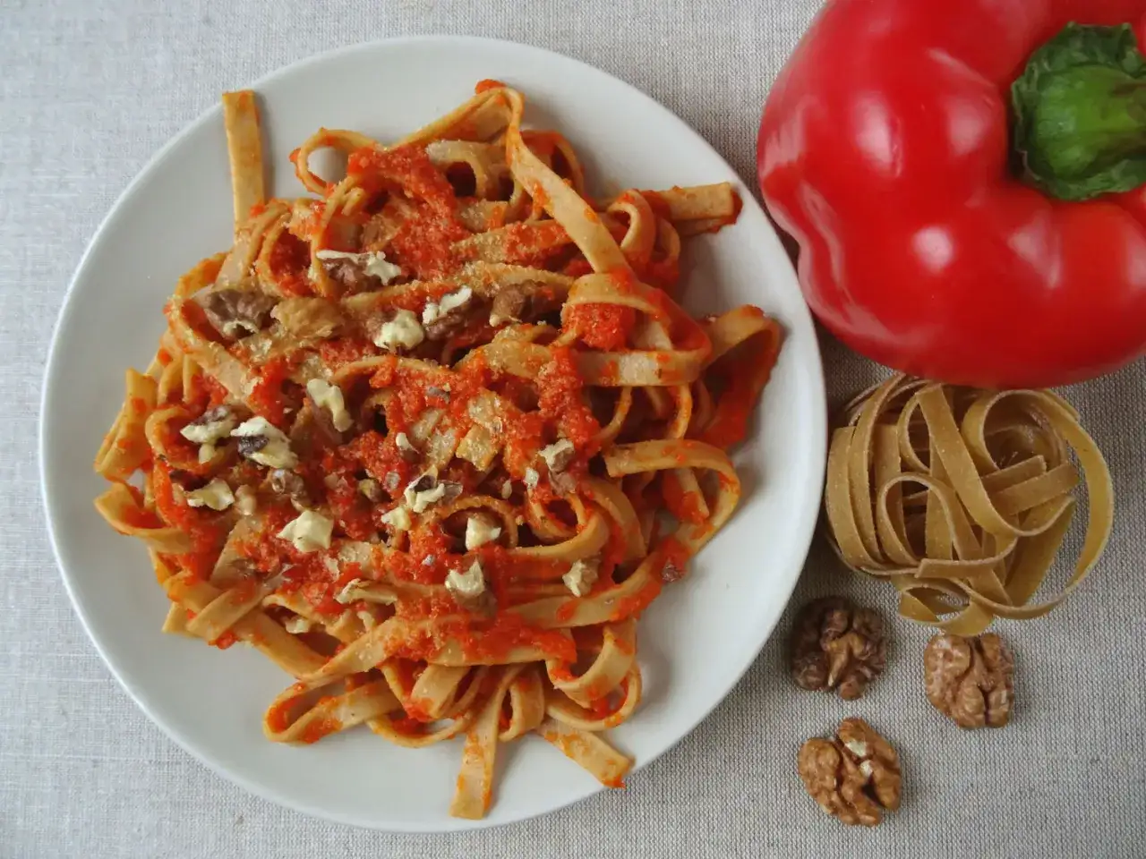 Makaron z sosem paprykowym i orzechami włoskimi, obok czerwona papryka i surowy makaron.