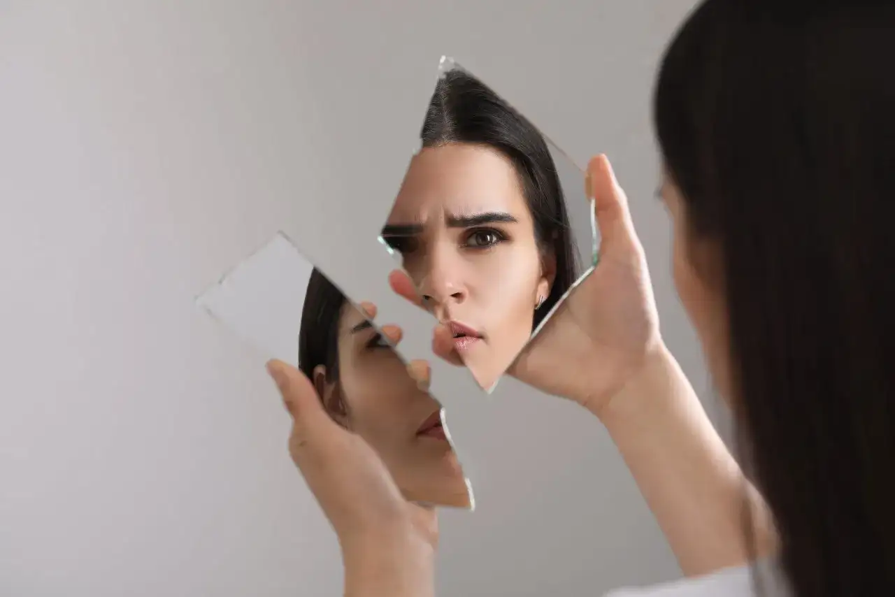 Kobieta patrzy na swoje odbicie w pękniętym lustrze, zastanawiając się, czym są kompleksy i jak wpływają na postrzeganie siebie.