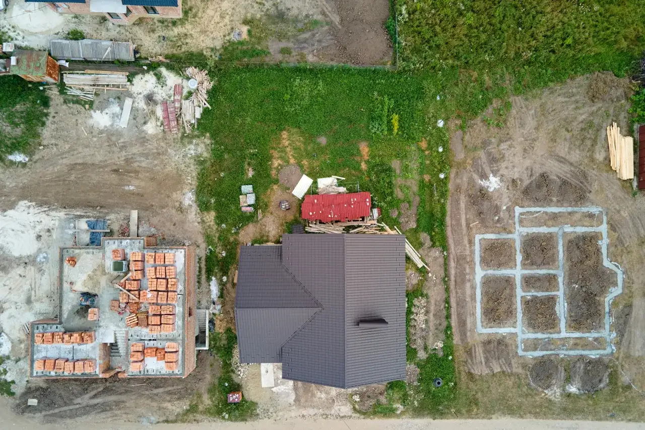 Widok z lotu ptaka na plac budowy z fundamentami, stosem cegieł i częściowo wybudowanym domem.