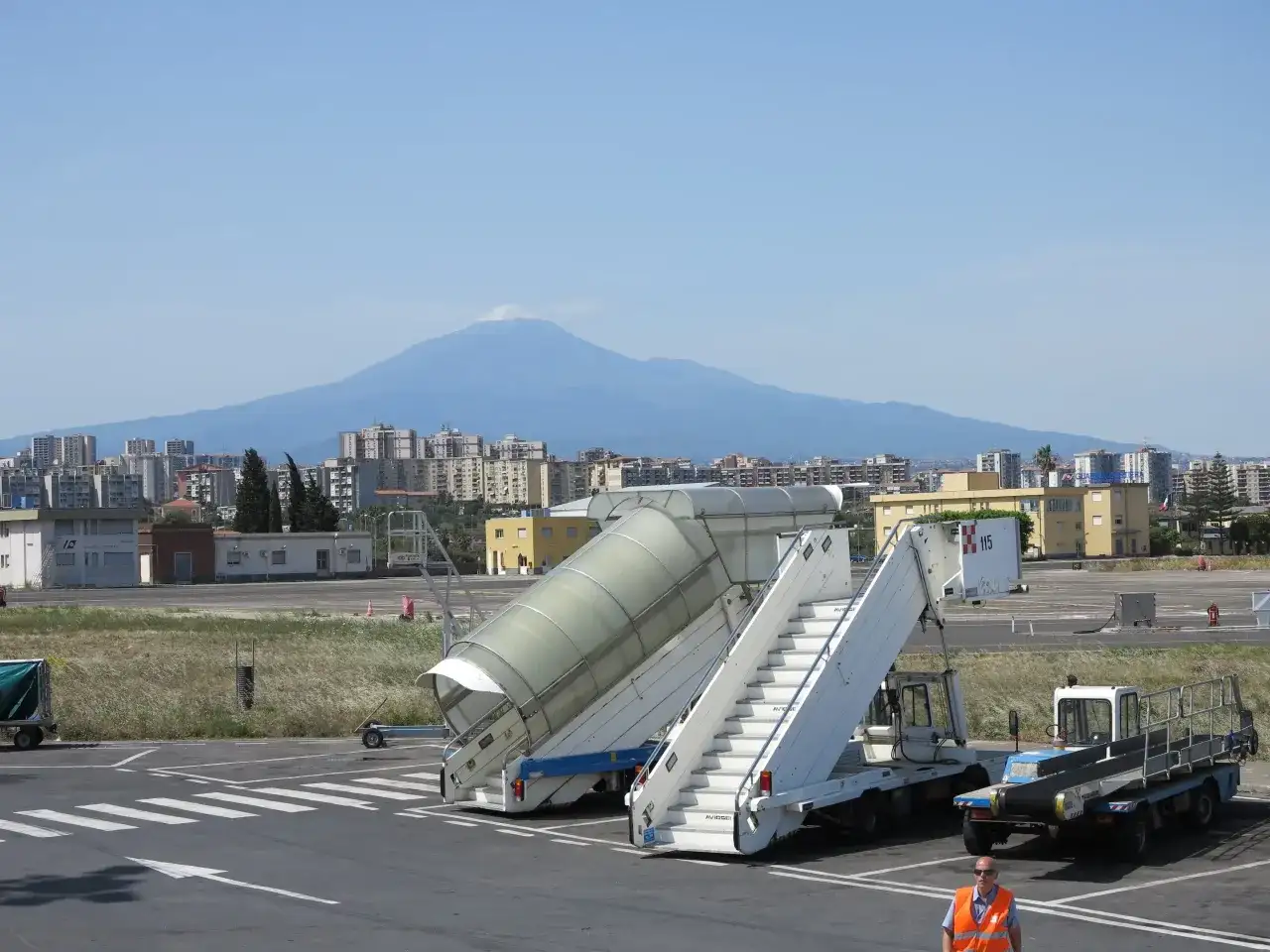 Port lotniczy Katania Fontanarossa z widokiem na Etnę. Schody lotnicze i pojazdy obsługi naziemnej na tle miasta i wulkanu.