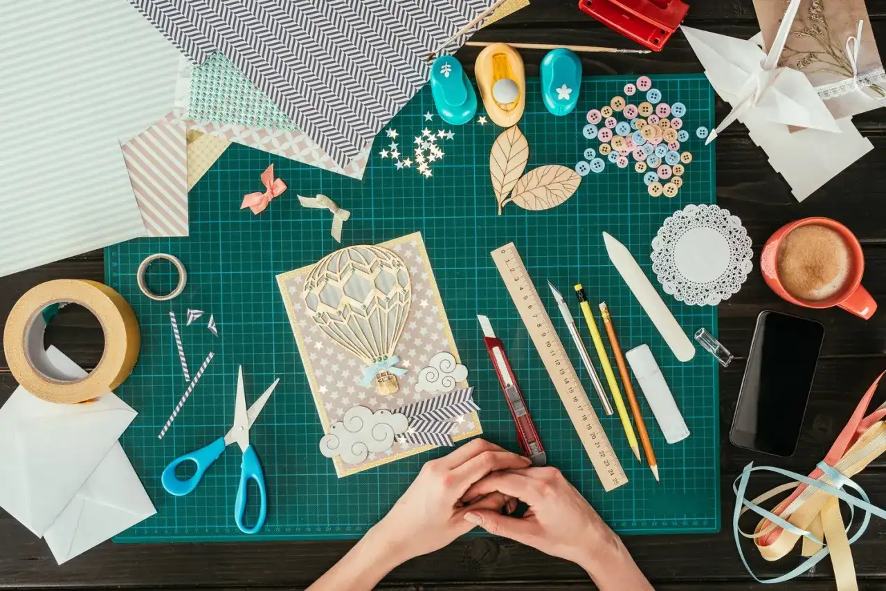 Ręce tworzą kartkę z balonem na gorące powietrze. Wokół materiały do scrapbookingu, taśma, nożyczki, guziki i inne akcesoria.