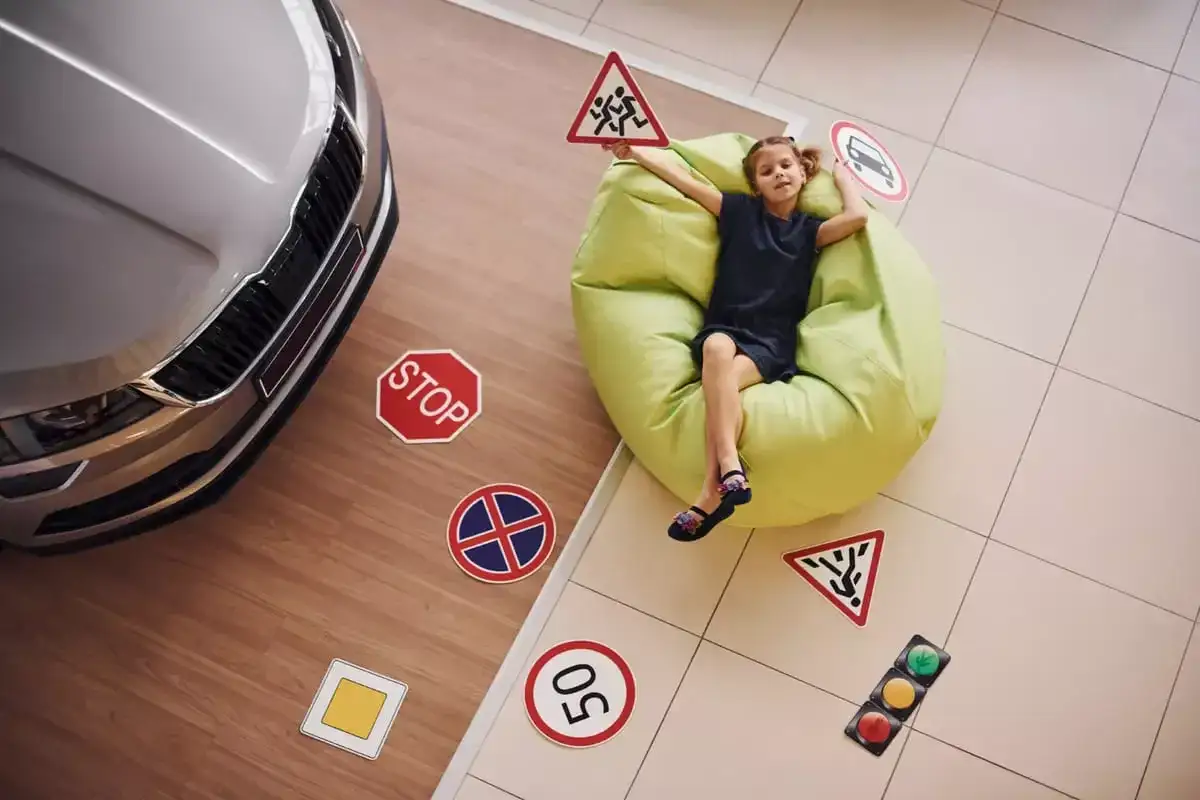Dziewczynka z tabliczkami drogowymi, jakby uczyła się, ile jest godzin jazdy na prawo jazdy. Obok samochód i znaki: stop, dzieci, ograniczenie prędkości.