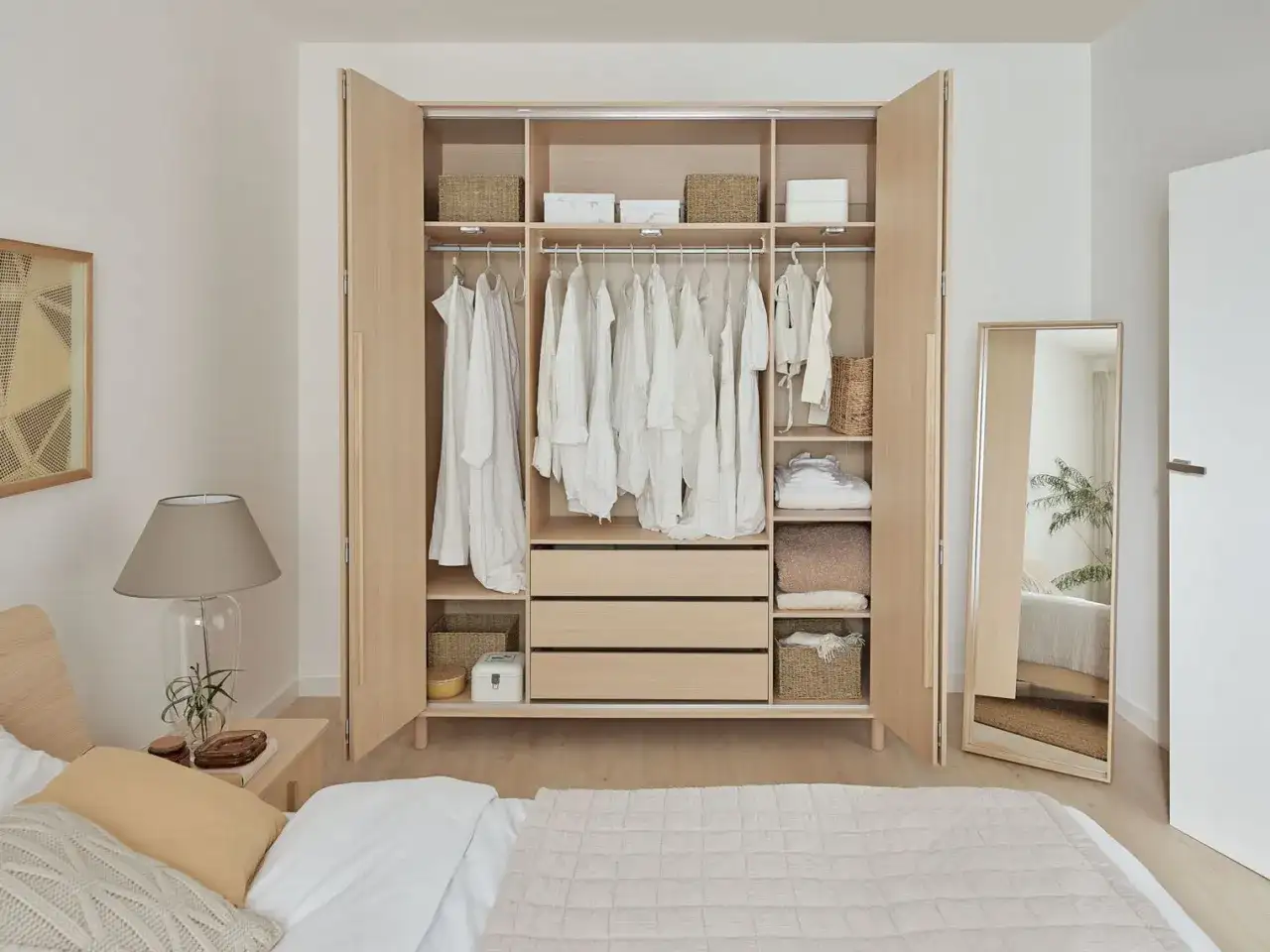Otwarta, jasna szafa z białymi ubraniami na wieszakach, półkami i szufladami. Obok lustro.