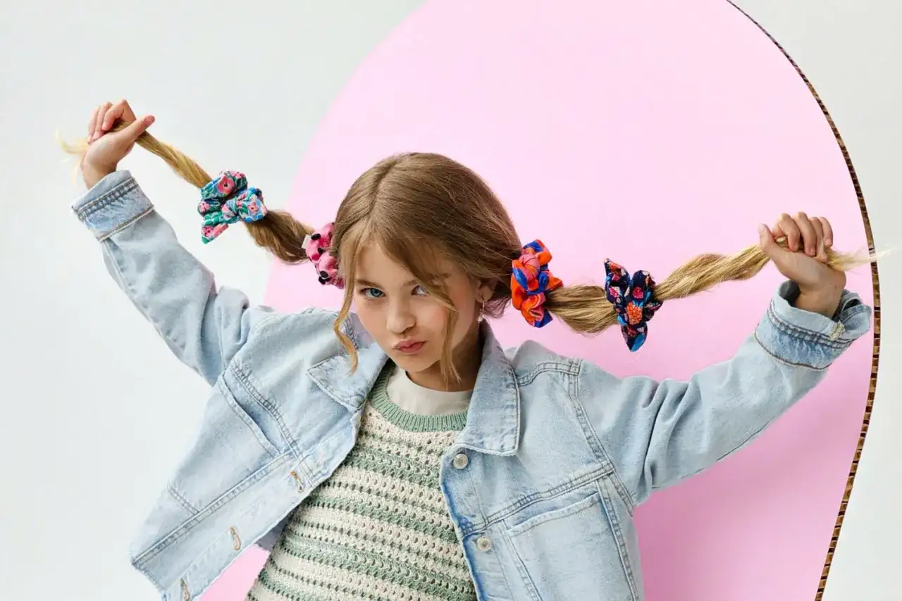 Dziewczynka z warkoczami ozdobionymi kolorowymi frotkami do włosów, w dżinsowej kurtce i swetrze.