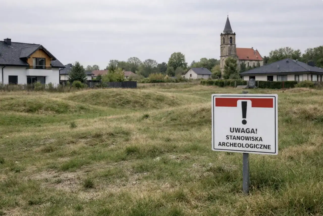 Znak "Uwaga! Stanowiska archeologiczne" na tle zabudowań i kościoła.