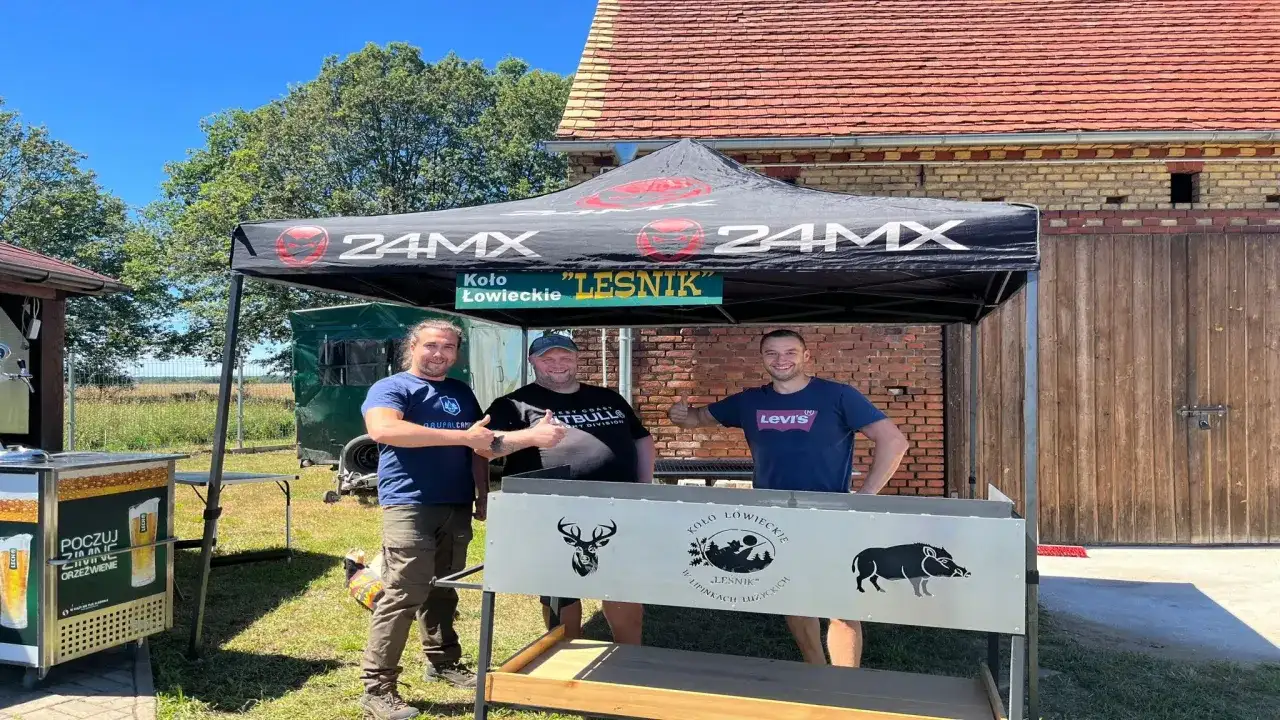 Trzech mężczyzn pod namiotem Koła Łowieckiego "Leśnik". Czy myśliwy może odsprzedać tuszę? Na grillu widnieją symbole zwierzyny.