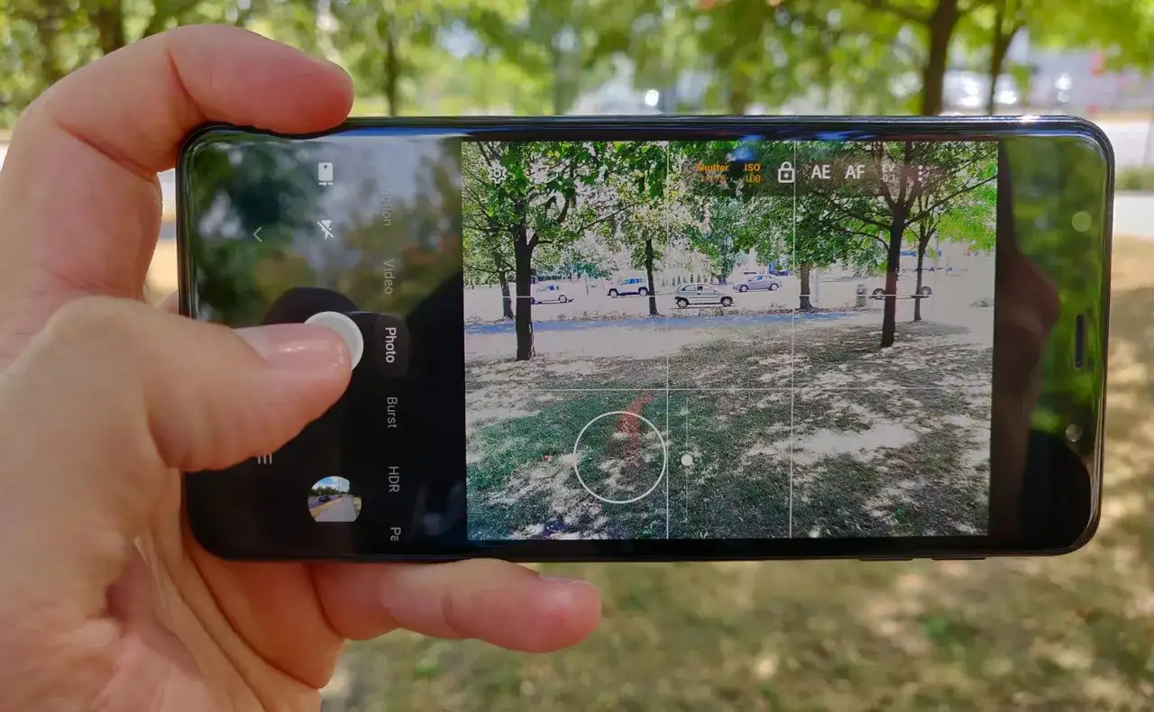 Dłoń trzyma smartfon, na ekranie którego widać interfejs aparatu. Aplikacja aparatu jest gotowa do zrobienia zdjęcia.