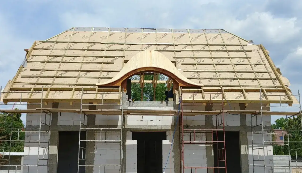 Budowa domu z charakterystycznym, drewnianym okno bawole oko na dachu. Widać rusztowanie i materiały budowlane.