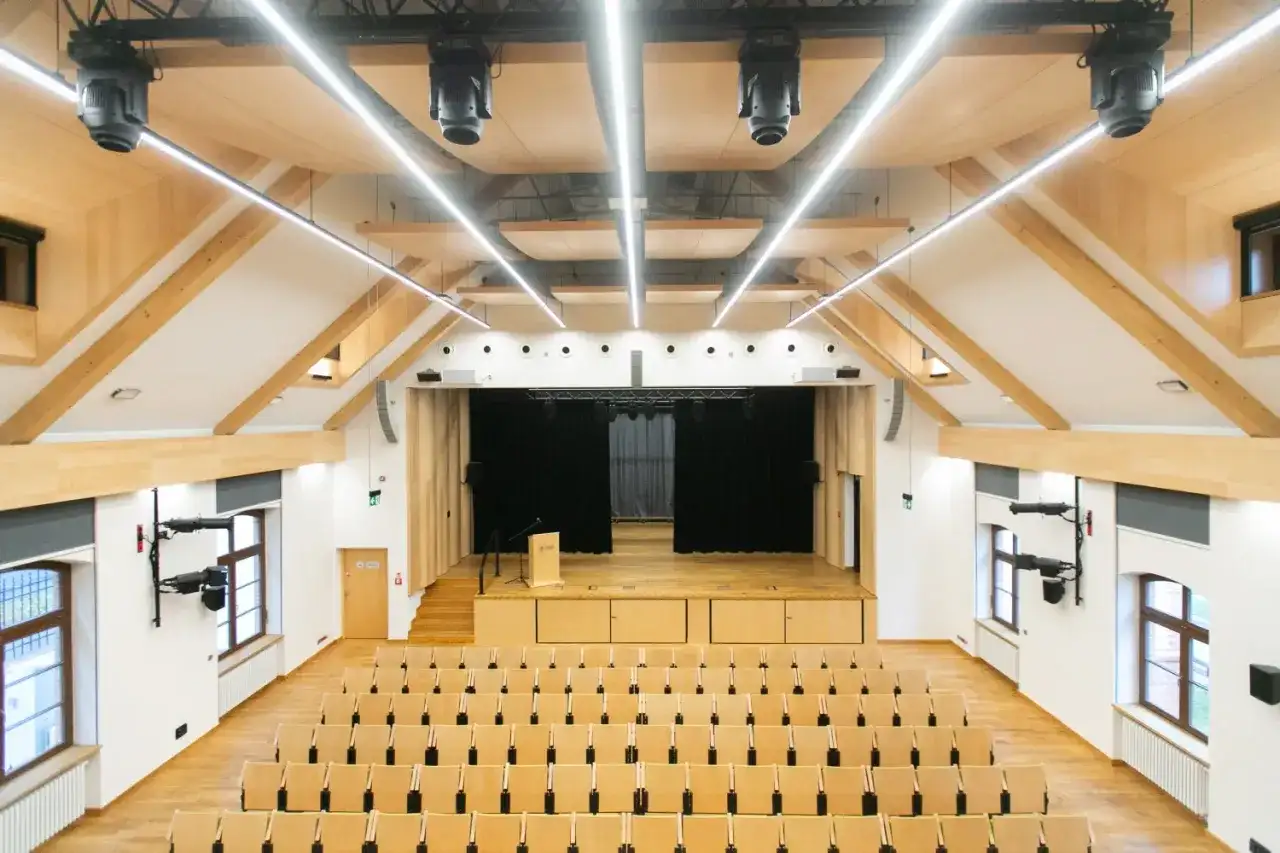 Nowa aula z drewnianymi elementami, sceną i rzędami krzeseł. Idealne miejsce na wykłady, koncerty i spotkania.