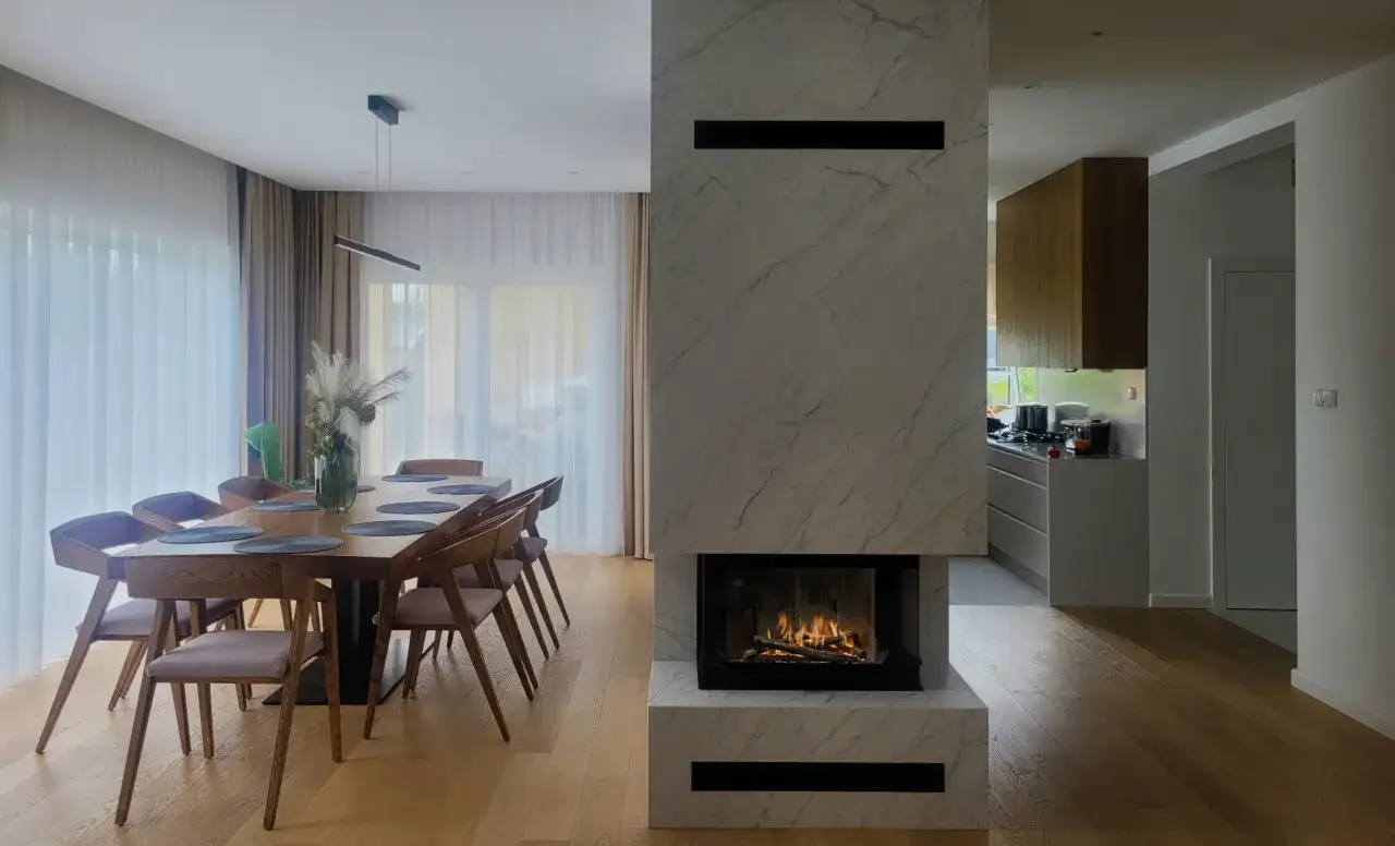 Nowoczesny kominek z marmuru, jak ozdobić komin w pokoju. Obok stół z krzesłami i kuchnia.