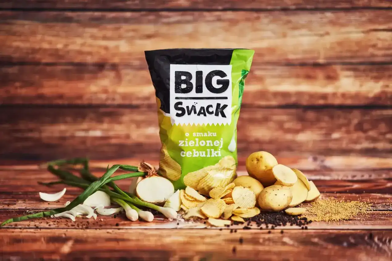 Reklamowa fotografia chipsów Big Snack o smaku zielonej cebulki, z ziemniakami i cebulką na drewnianym tle.