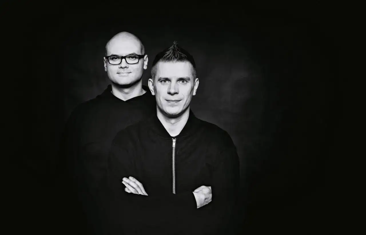 Dwóch mężczyzn w czarnych bluzach na ciemnym tle. Jeden z nich ma okulary, drugi irokeza. Wyglądają jakby właśnie wylądowali po locie dzika.