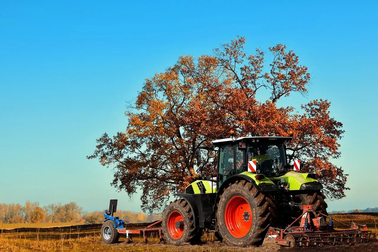 Zielony traktor z pługiem pod jesiennym drzewem. Może posłużyć jako inspiracja do wypełnienia wniosku o odrolnienie działki wzór.