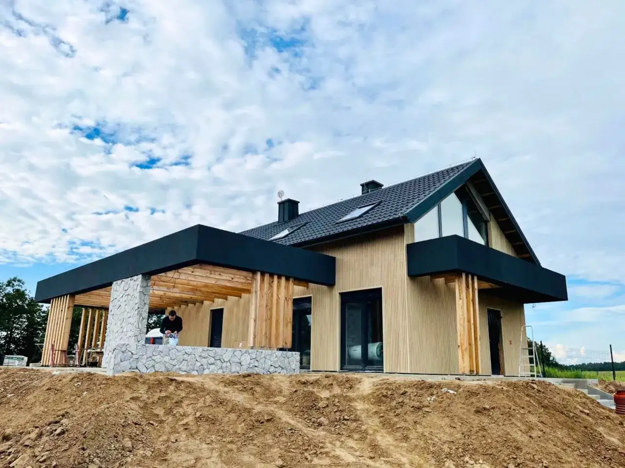 Nowoczesny dom w stanie deweloperskim, z drewnianą elewacją i kamiennym cokołem. Widać prace wykończeniowe na tarasie.