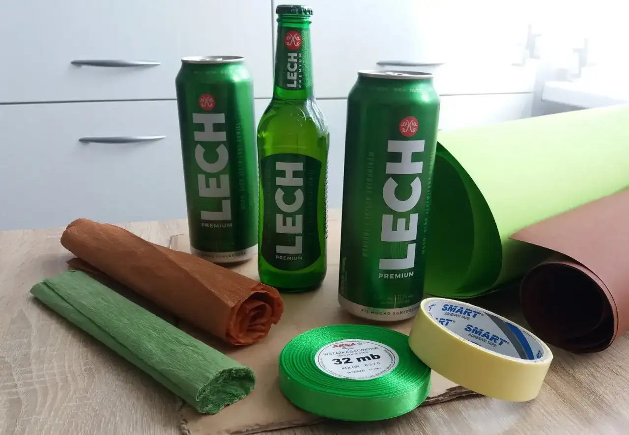 Puszki i butelka piwa Lech, rolki papieru, wstążka i taśma klejąca na drewnianym stole.