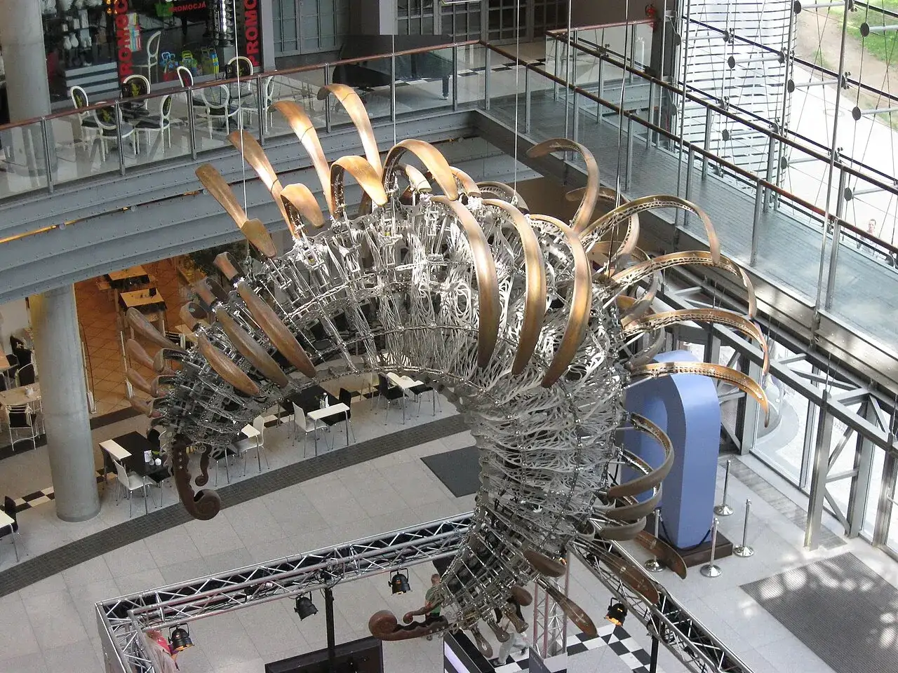 Duża, metalowa rzeźba przypominająca szkielet dinozaura w centrum handlowym.