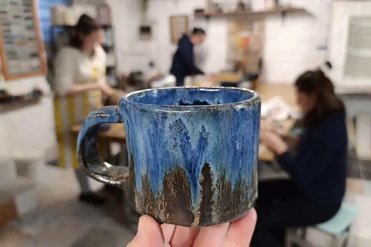 Ręcznie robiony, niebiesko-brązowy kubek ceramiczny trzymany w dłoni, w tle pracownia ceramiczna.