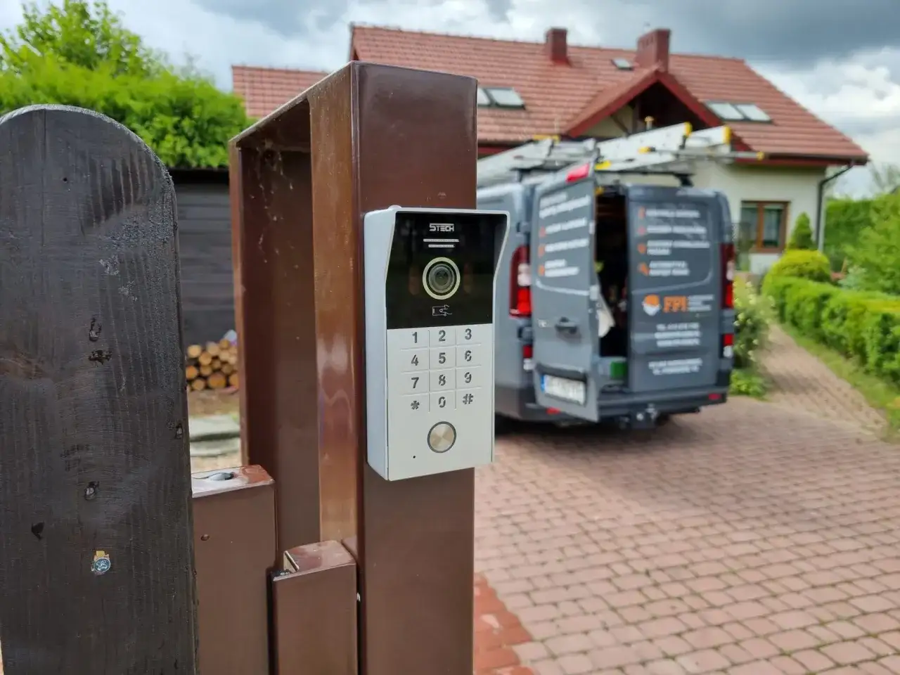 Nowy domofon na furtce. Zastanawiasz się, ile kosztuje montaż domofonu w domu jednorodzinnym? W tle widać dom i samochód firmy instalacyjnej.