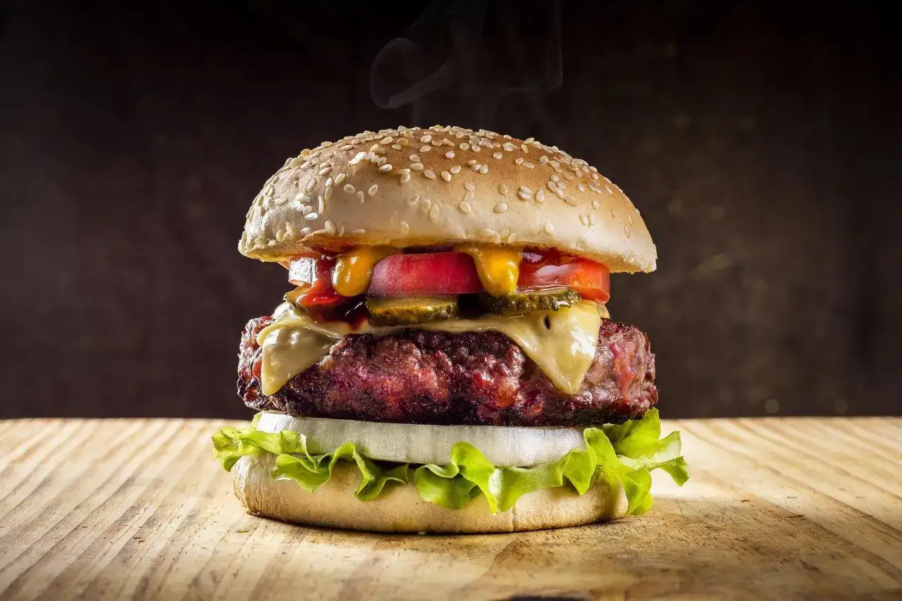 Parujący, soczysty burger z serem, warzywami i sosami na drewnianym blacie.