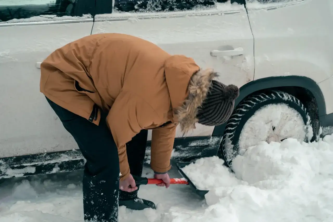 Osoba w zimowej kurtce odśnieża samochód uwięziony w zaspie śnieżnej.