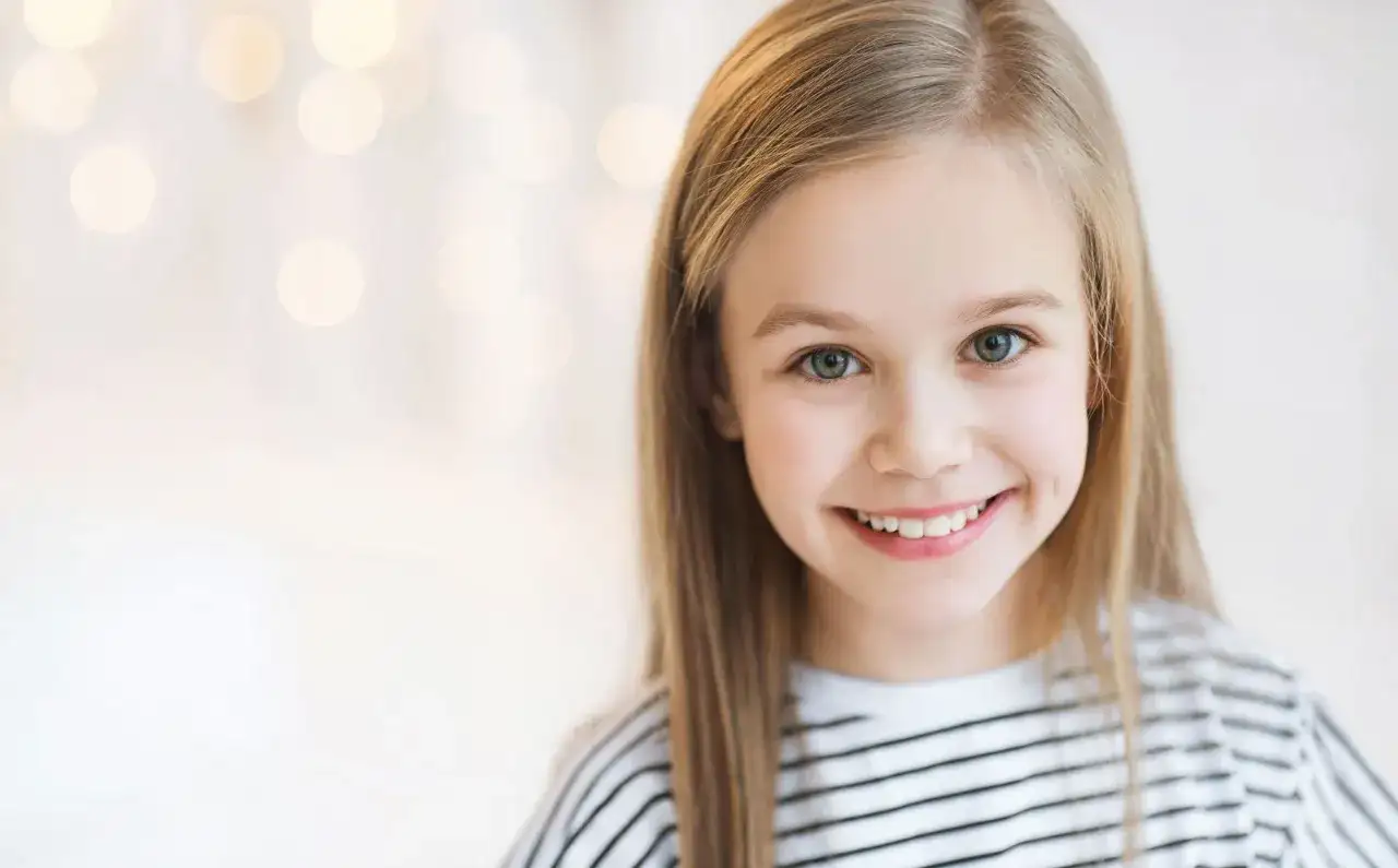 Uśmiechnięta dziewczynka o blond włosach, jakby inspirowana nowoczesnymi imionami dla dziewczyn, z rozmazanym tłem pełnym świateł.