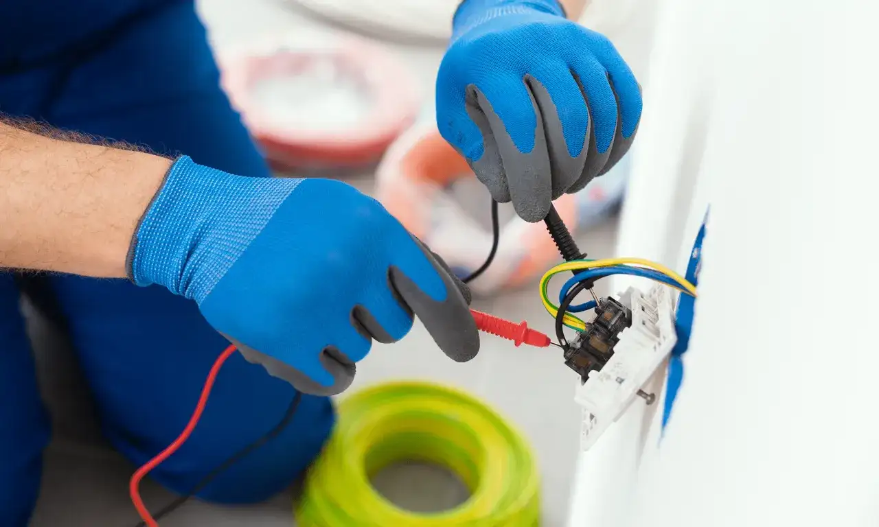 Elektryk w niebieskich rękawicach sprawdza połączenia przewodów. W tle widać rolki kabli. To instrukcja, jak podłączyć gniazdko bez uziemienia.