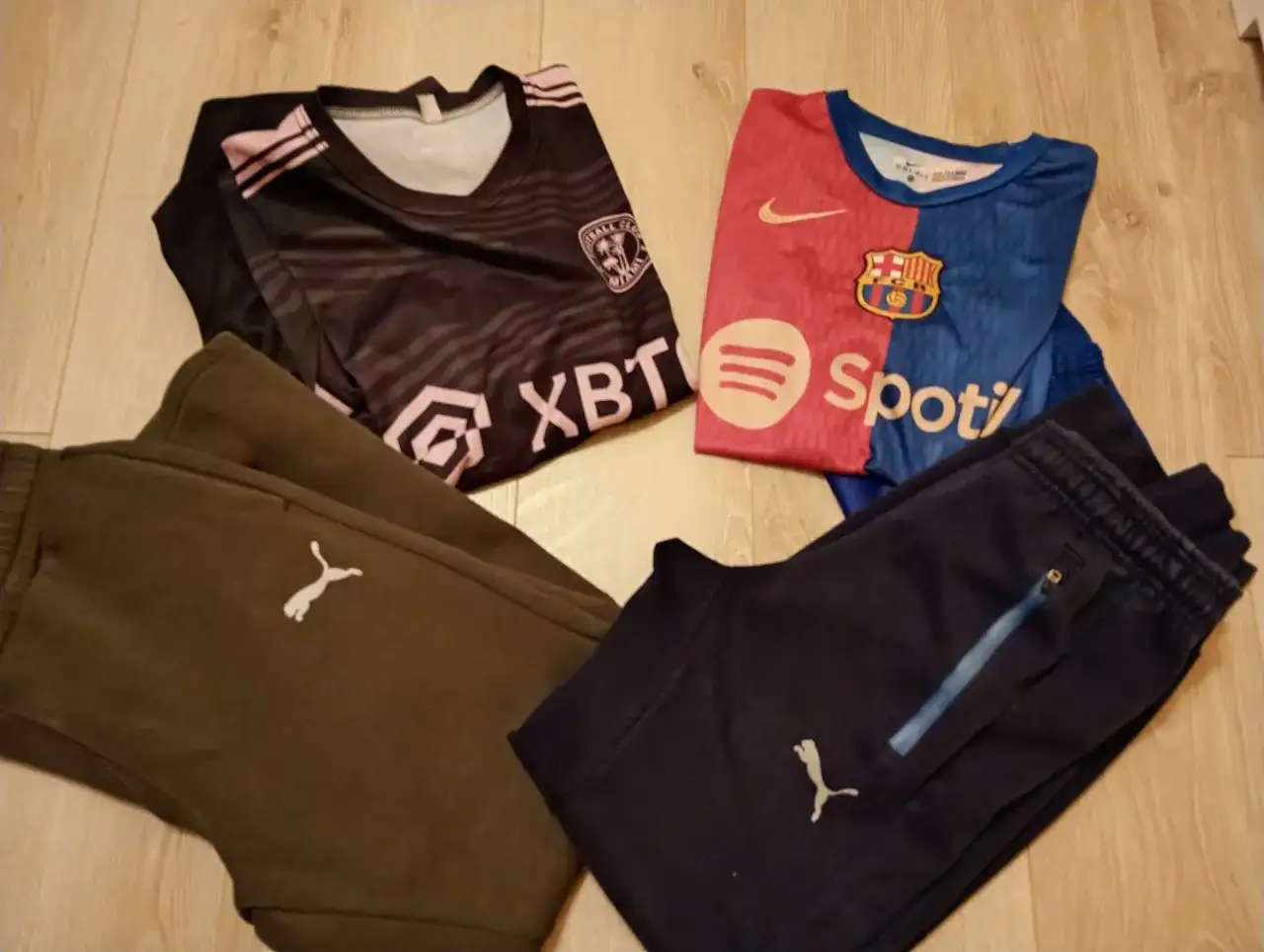 Dwa komplety odzieży piłkarskiej dla chłopca: czarna koszulka z różowymi pasami i herbem, niebiesko-czerwona koszulka FC Barcelony, brązowe i granatowe spodnie piłkarskie chłopięce z logo Pumy.
