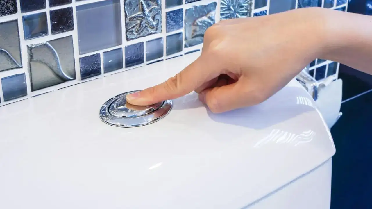 Dłoń naciska chromowany przycisk spłuczki WC na białej ceramice, w tle mozaika.