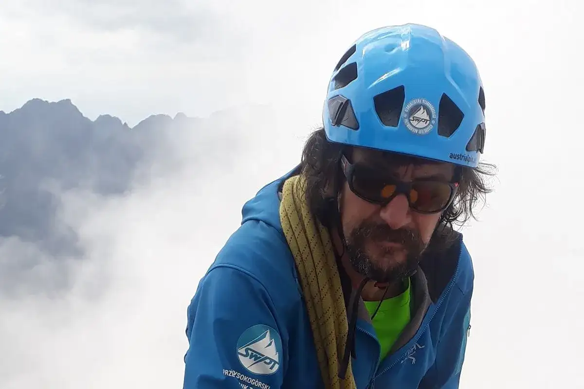 Mężczyzna w kasku i okularach przeciwsłonecznych, z liną wspinaczkową na szyi, na tle mglistych szczytów. Perć w Tatrach.
