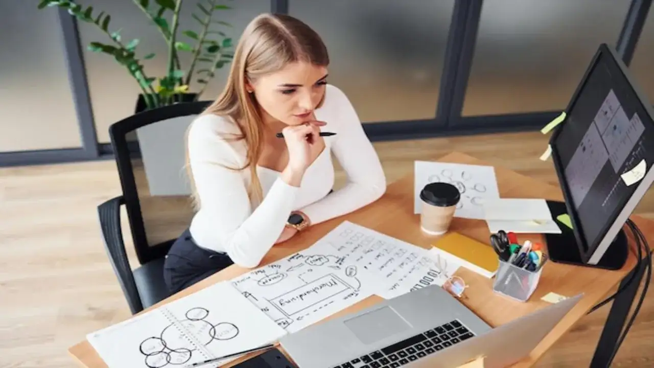 Młoda kobieta analizuje dokumenty, zastanawiając się, ile zarabia projektant mody. Na biurku laptop, szkice i kawa.