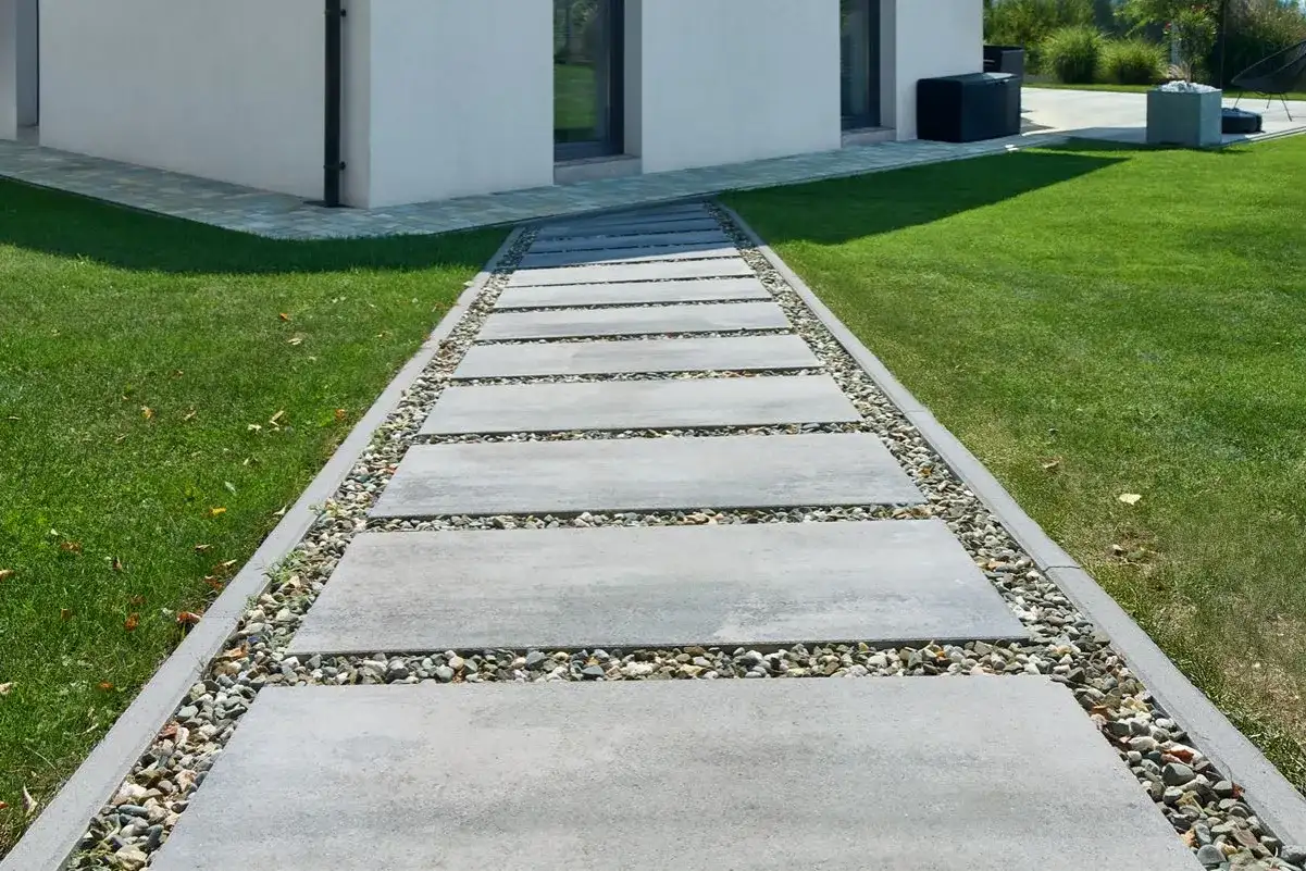Ścieżka z prostokątnych płyt betonowych i kamieni ozdobnych obok trawnika i białego domu.