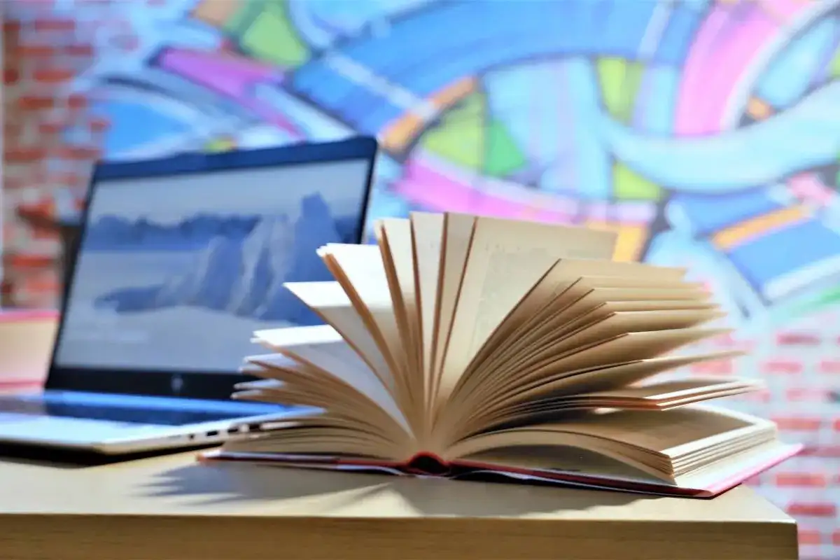 Otwarta książka na biurku obok laptopa, w tle kolorowe graffiti.