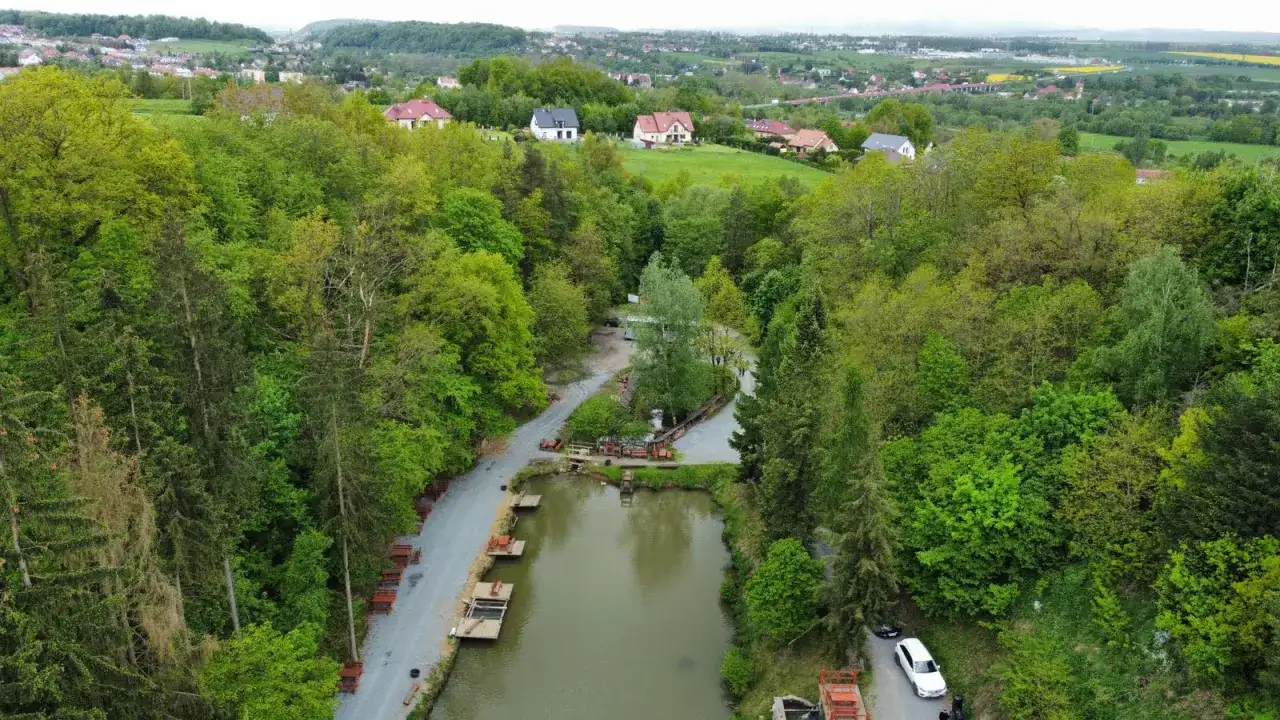Widok z lotu ptaka na łowisko pstrągowe otoczone zielonymi drzewami, z domami w tle.
